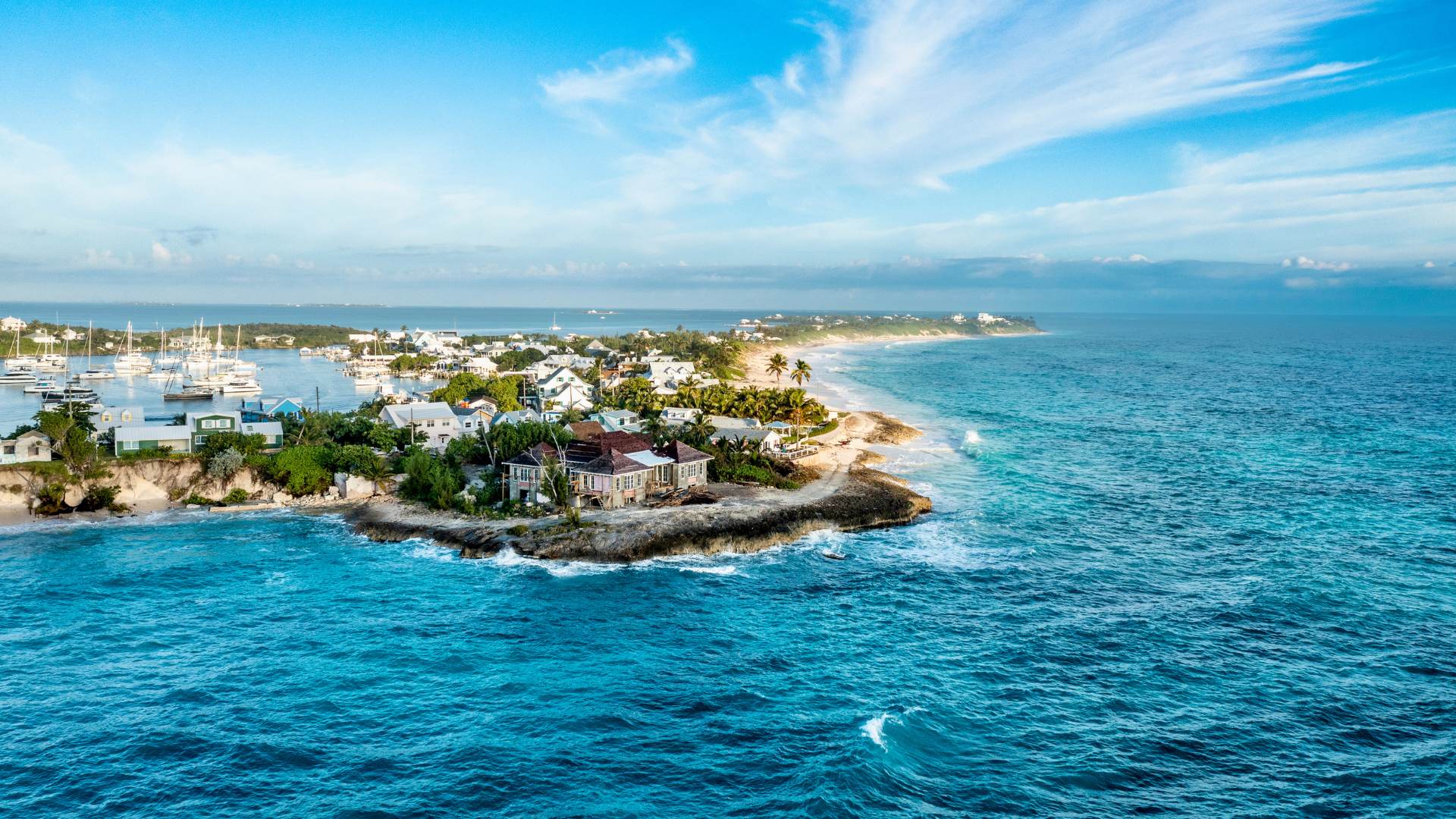 Aerial of Hope Town's harbor point on Elbow Cay — yachts moored inside, cottages along the shore, surf on the ocean side