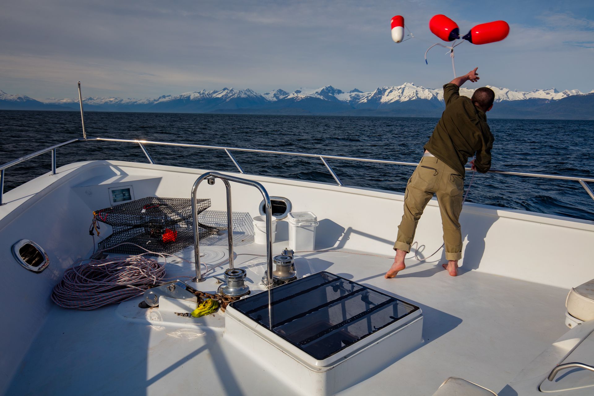 A crew member sends a crab-pot buoy off the bow, a dramatic snow-mountain panorama behind — the slower, scenery-forward register the trip settles into on the run down toward Baranof.