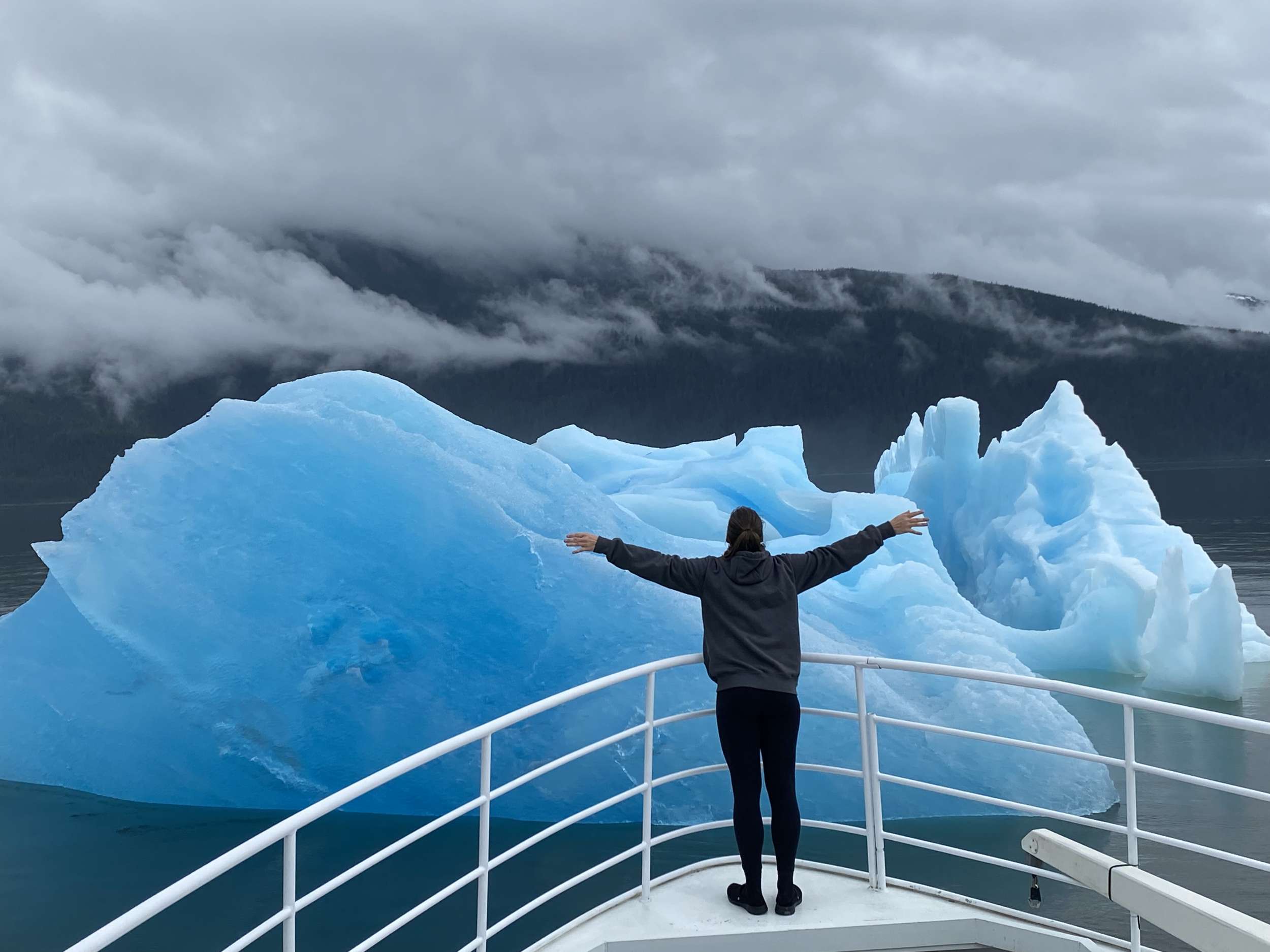 Guest at the bow rail before towering blue berg ice, fog draped on the mountains behind