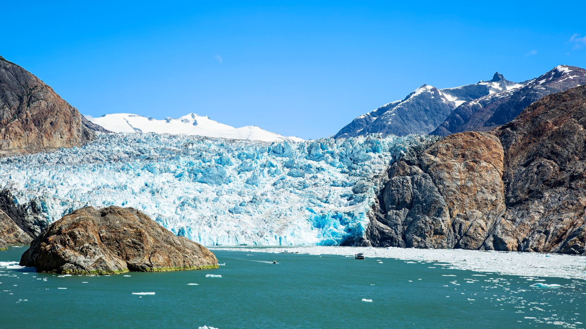 The blue calving face of a tidewater glacier filling the head of a fjord, small boats dwarfed beneath it