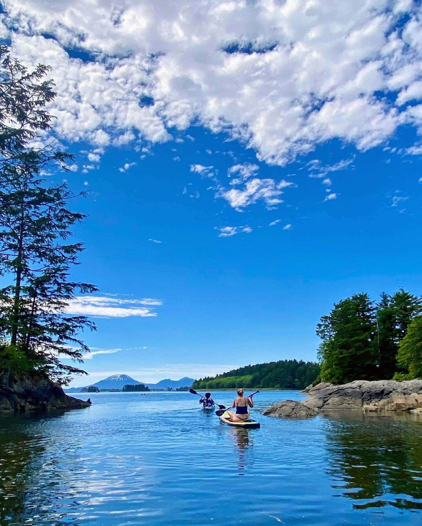 A kayaker and paddleboarder threading a rocky spruce cove on Sitka Sound, a volcano cone on the horizon