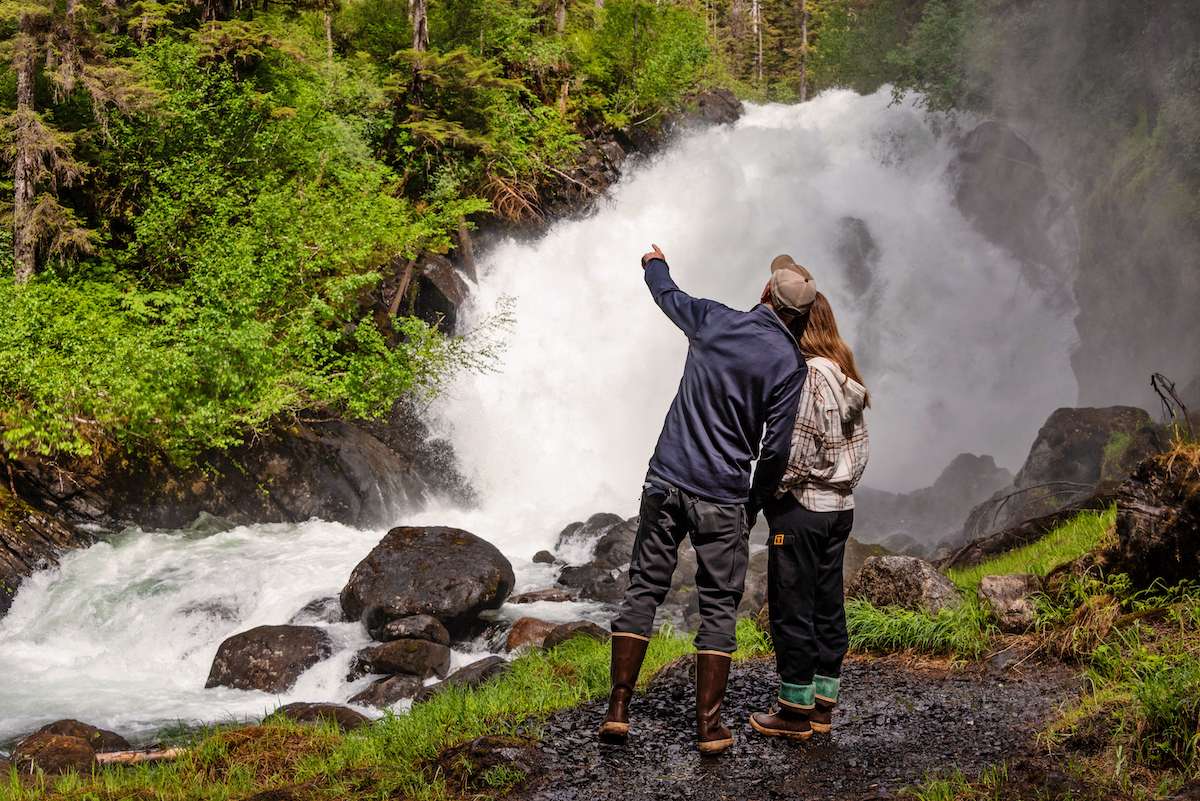 A couple in rain gear at the base of a granite-walled waterfall — the hanging-valley cascades this stretch of coastline is known for.