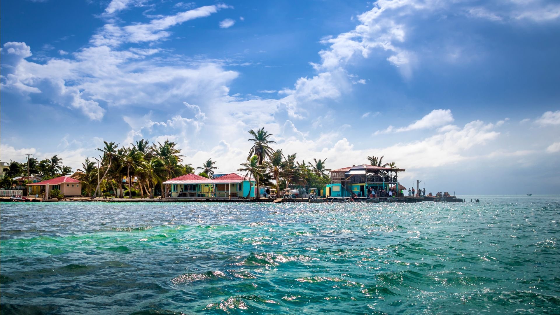 Caye Caulker waterfront with palms and colorful stilted island buildings over clear turquoise water.