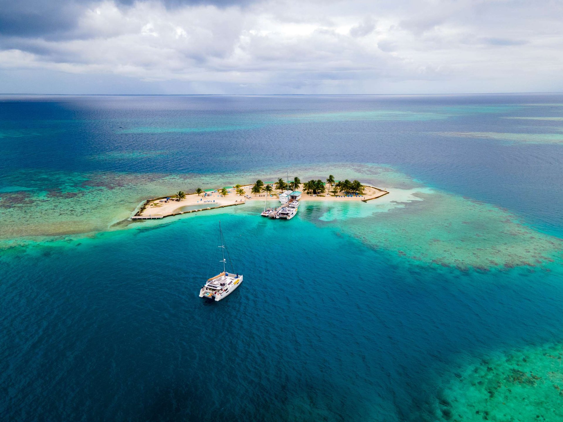 A catamaran at anchor off a small palm-covered caye with a beach deck and umbrellas.
