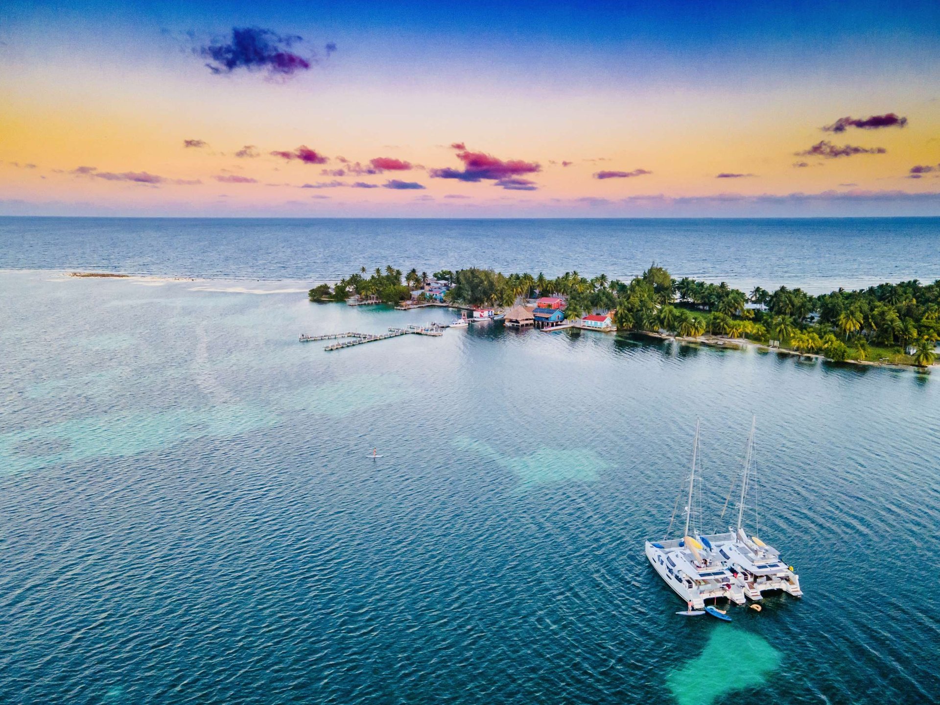Pastel sunset over a catamaran at anchor off a palm-covered caye in Belize.