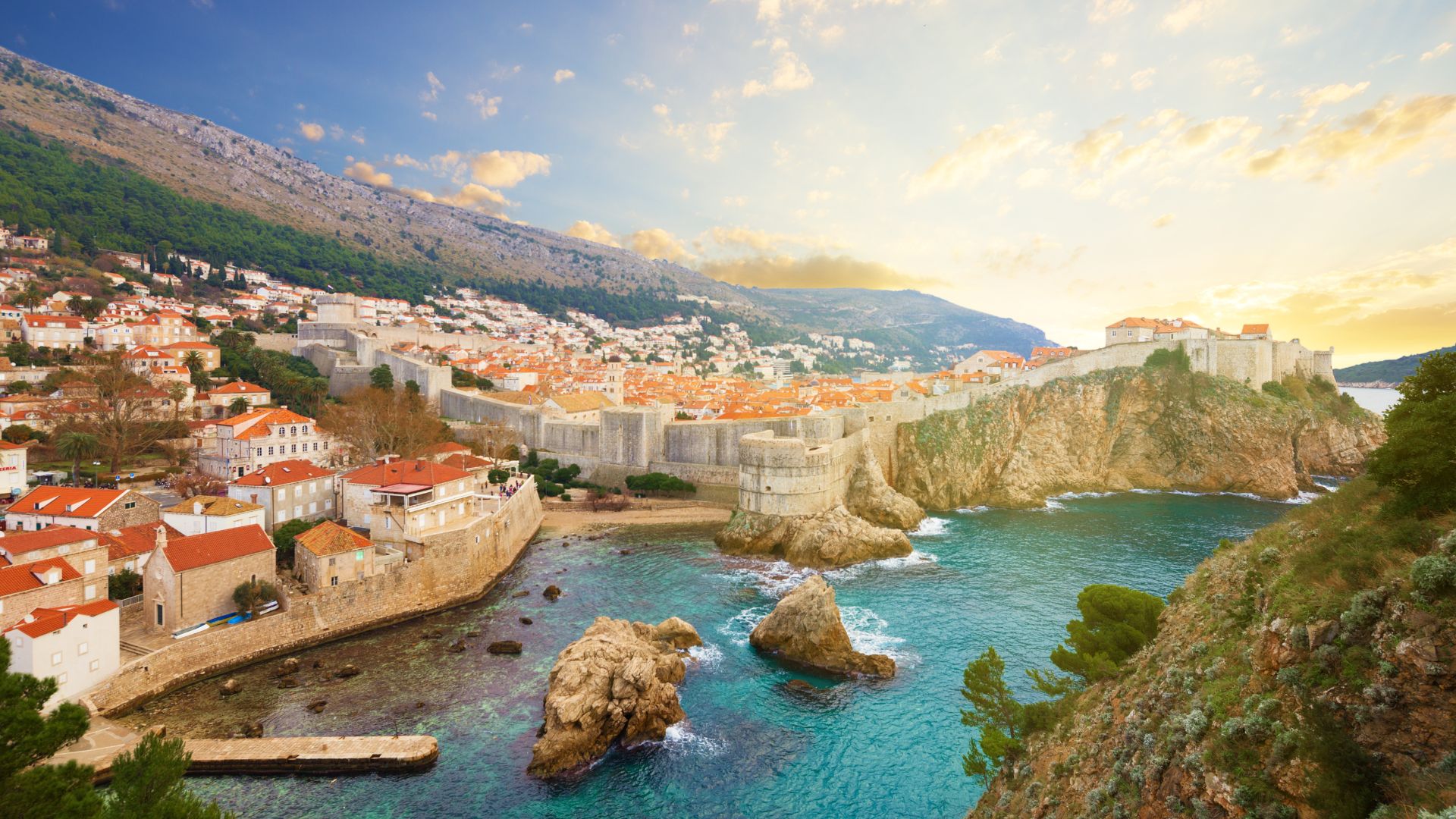 Dubrovnik from the Lovrijenac fortress side at golden hour — city walls in profile, sea rocks in the foreground.