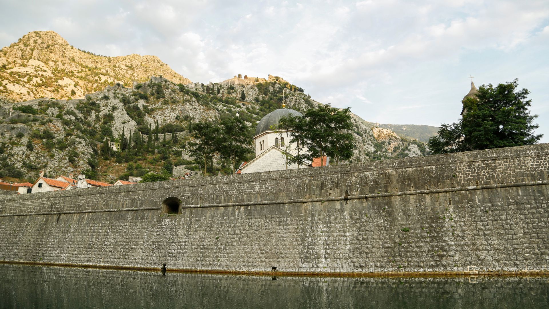 Kotor's medieval curtain wall along the Škurda waterway — small grey-domed chapel above the wall, the Lovćen mountain wall behind