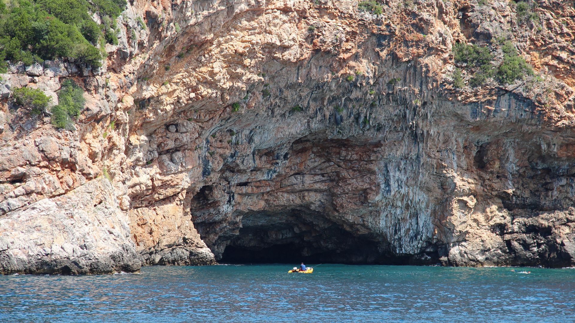 Tender approaching the Blue Cave (Plava Špilja) on the Luštica peninsula — a small inflatable at the dark cave mouth in the tan limestone cliff.