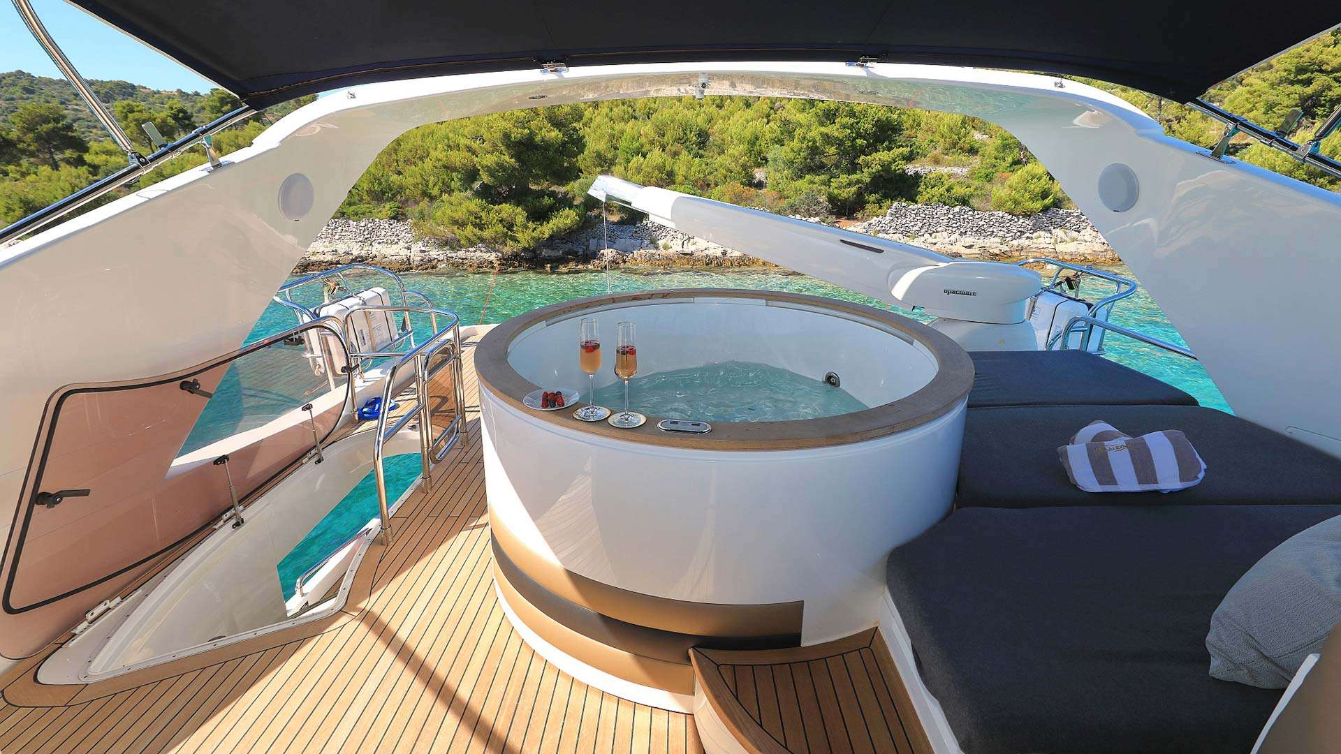 Aft-deck jacuzzi off a motor yacht's swim platform — two glasses of champagne, a pine-shore anchorage behind