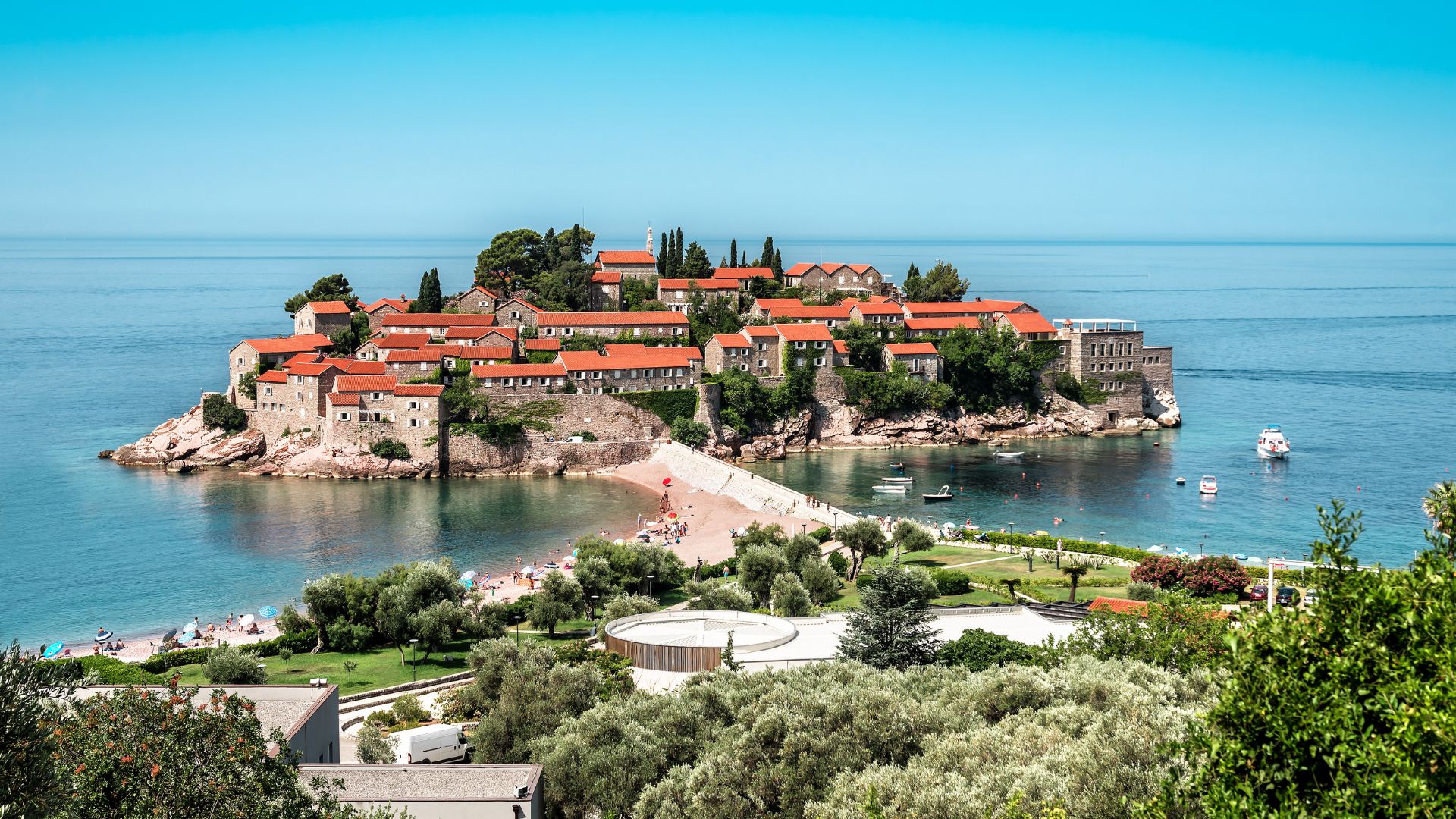 Sveti Stefan from the cliff above — fortified island, terracotta roofs, pink-pebble causeway to the mainland