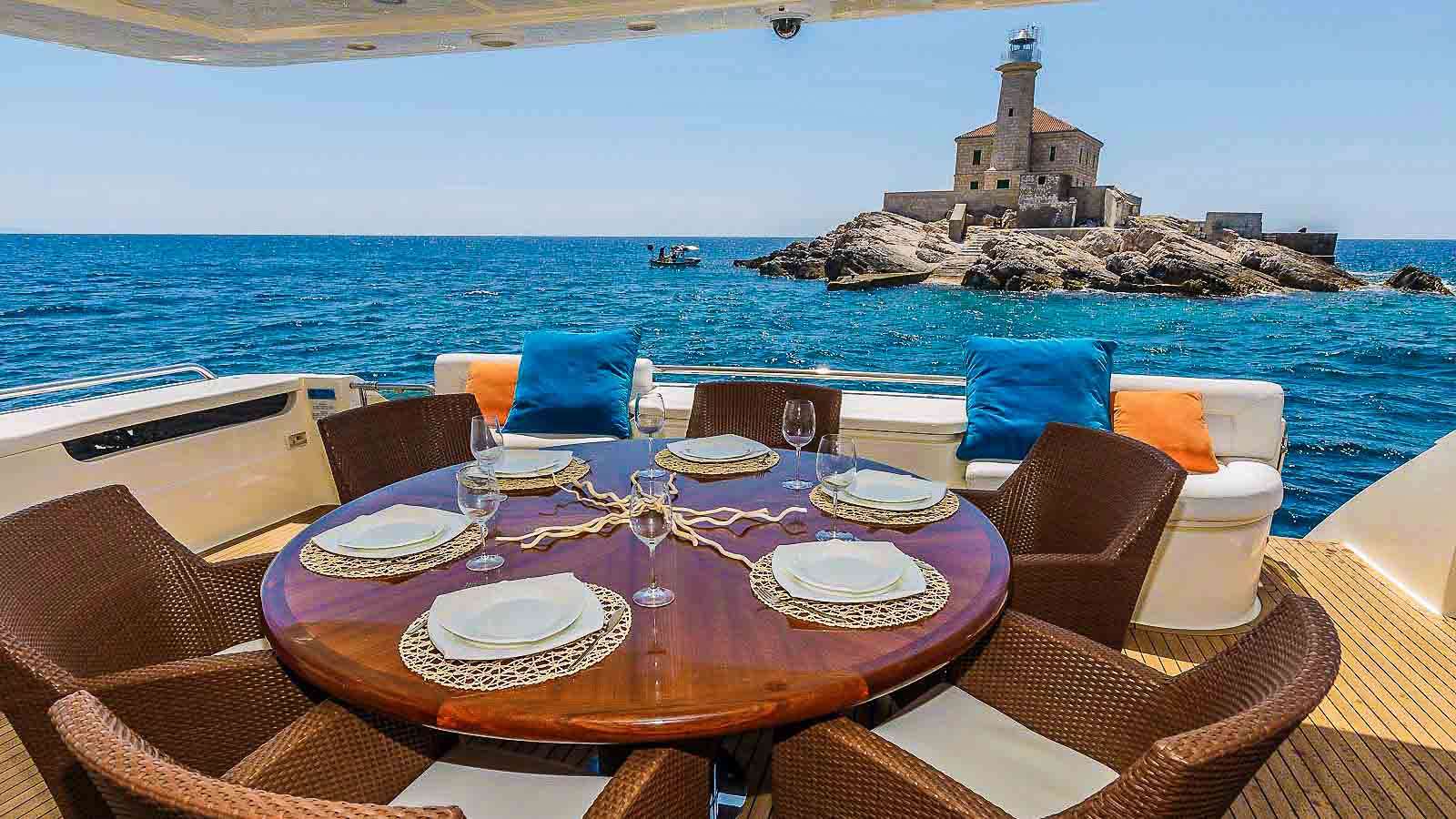 Round wooden dining table set on a crewed yacht aft deck — six places, evening light off a small rocky islet astern