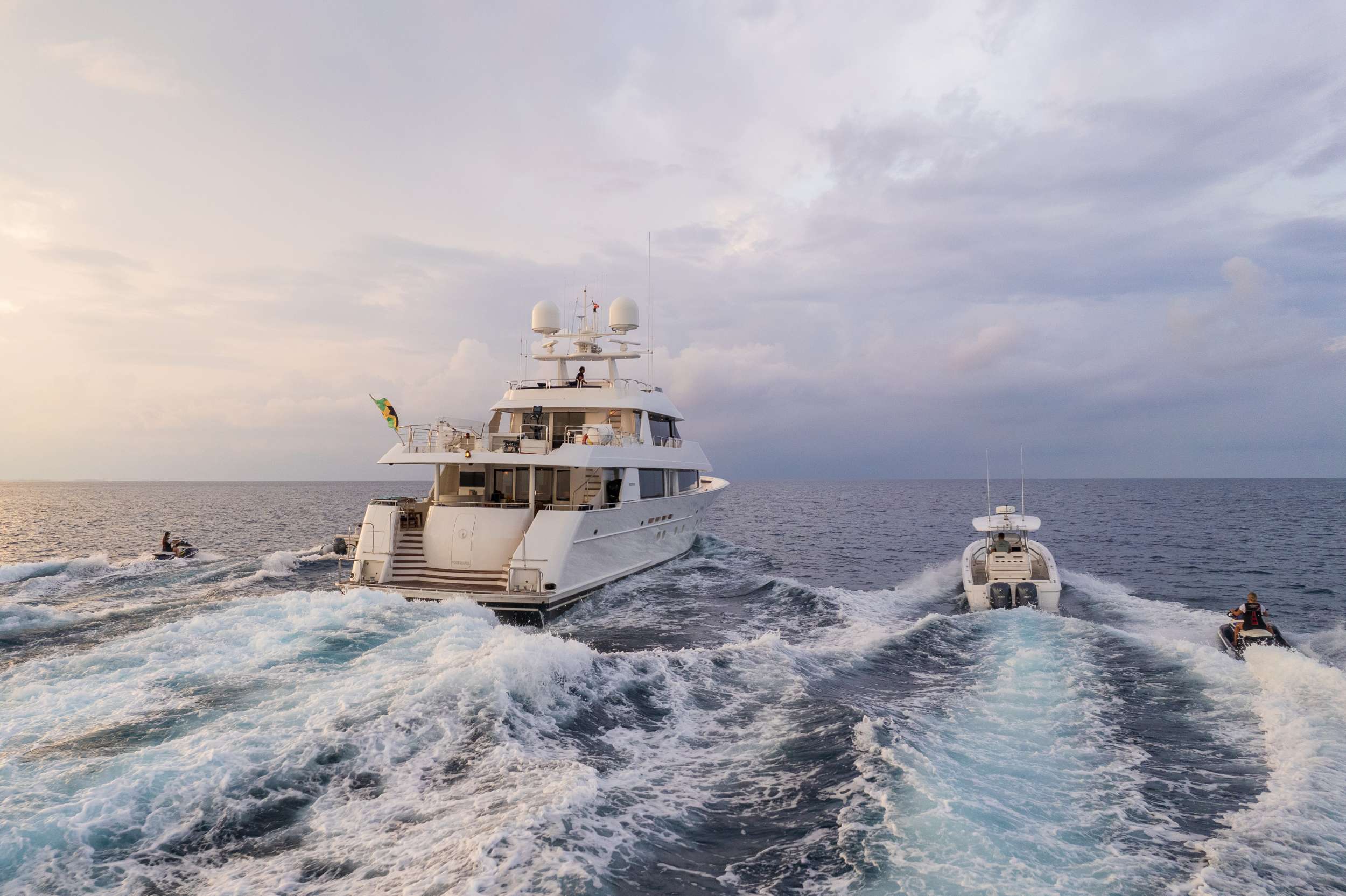 Crewed motor yacht under way at dusk with tender and jet skis running alongside