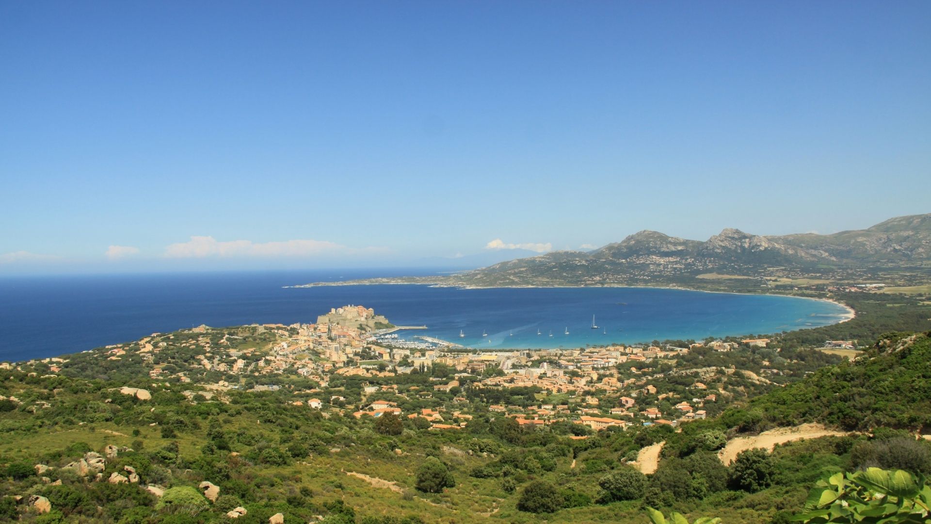 Calvi's bay holds a five-mile sand beach, with the Genoese citadel on the western headland and the snow-capped Monte Cinto range on the horizon inland.