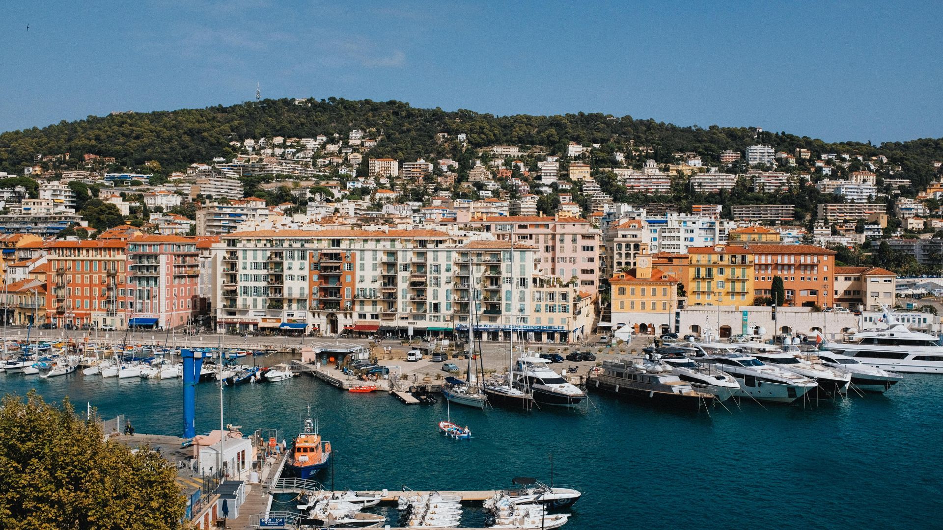 Cannes harbor — megayachts side-tied along the quay during the season, the Croisette running along the seafront, and the Lerins Islands visible across the bay.