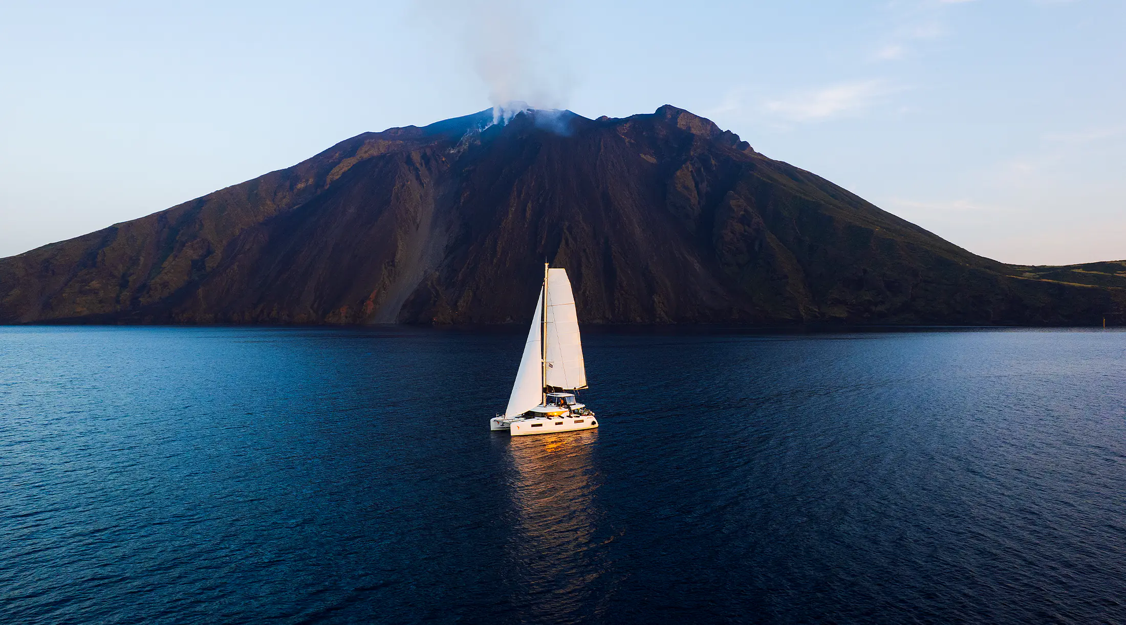 Crewed sailing catamaran passing the smoking cone of Stromboli at dusk — the marquee scene of an Aeolian Islands charter week.