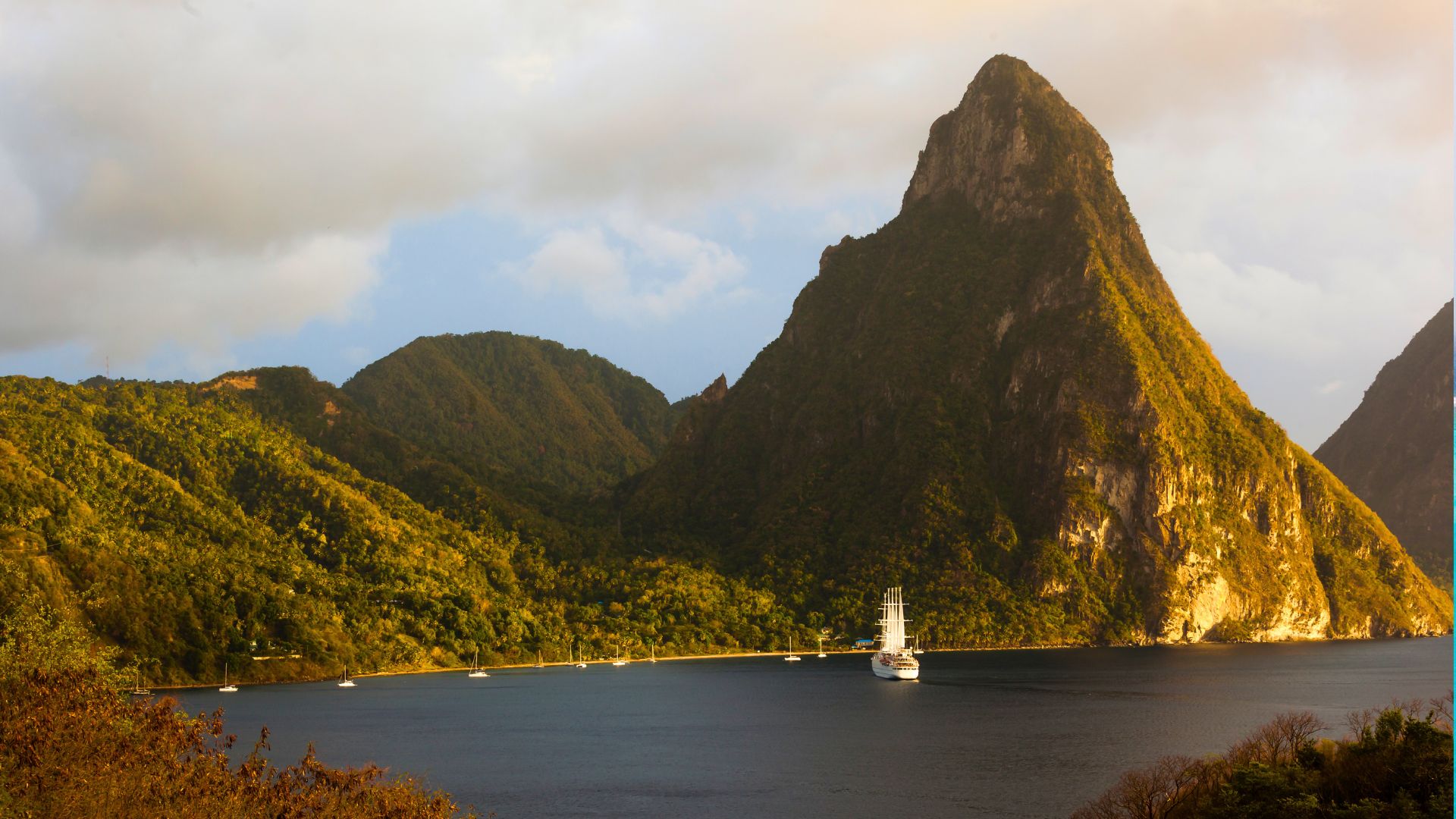 A Piton lit gold by late-afternoon sun rising above Soufrière Bay, St. Lucia, with a tall ship and yachts anchored in the water below.