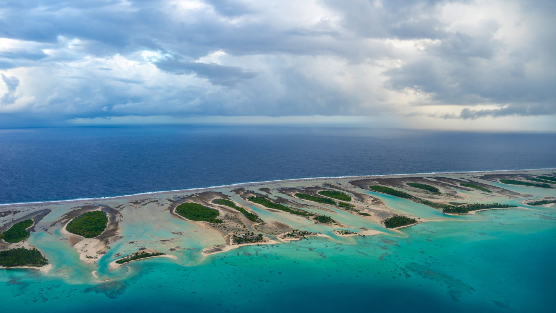 The outer reef edge of a Tuamotus atoll — palm-covered motus on the inside, the open Pacific breaking on the reef on the outside, and the line between blue and turquoise that defines the chain from above.