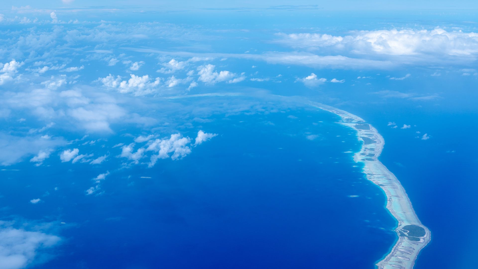High-altitude aerial of a Tuamotus coral atoll—a thin curving reef line wrapping a vast turquoise lagoon, surrounded by deep blue Pacific Ocean.