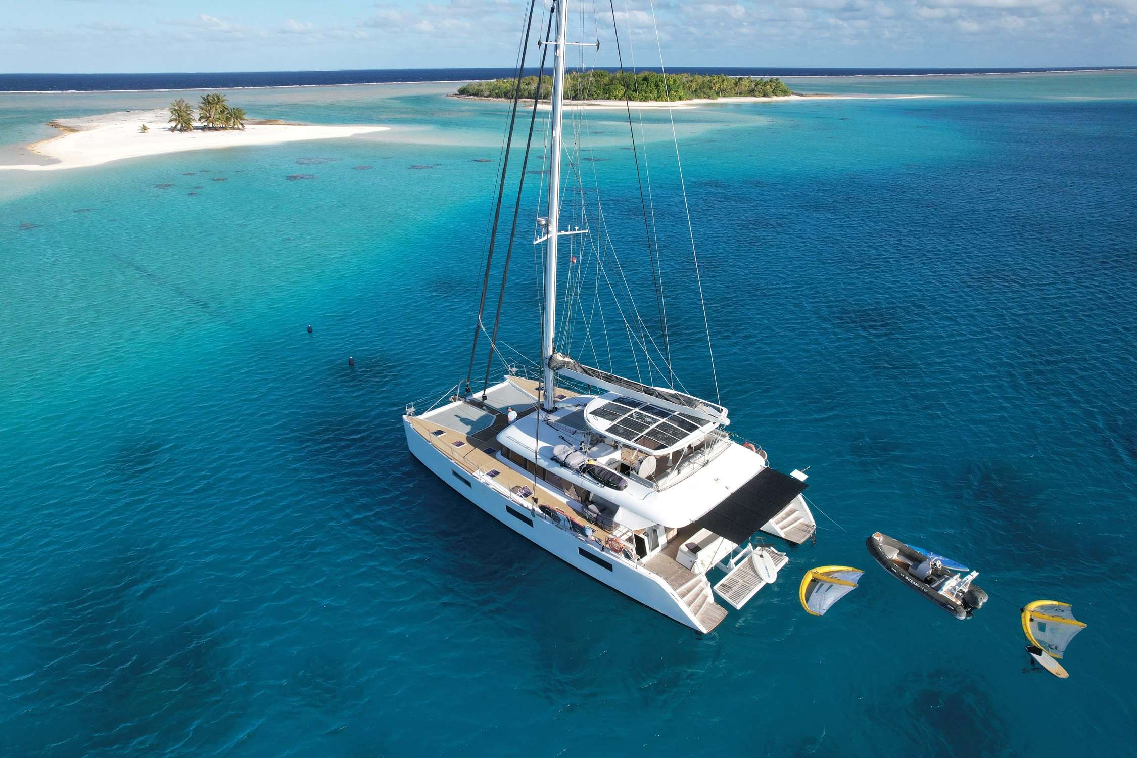 A crewed sailing catamaran at anchor in a Tuamotus lagoon with paddleboards and water toys deployed off the swim platform, palm-covered motu in the background.
